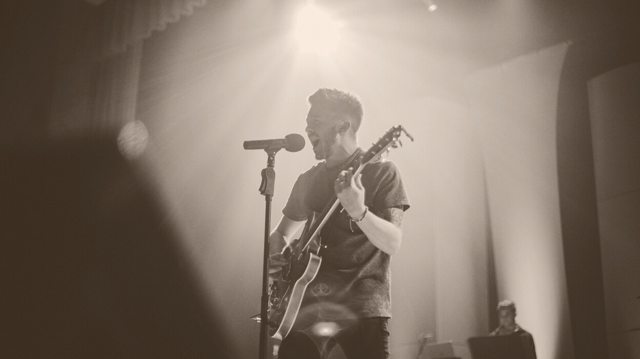 Man Playing Guitar Standing on Stage Singing in Front of Standing Microphone